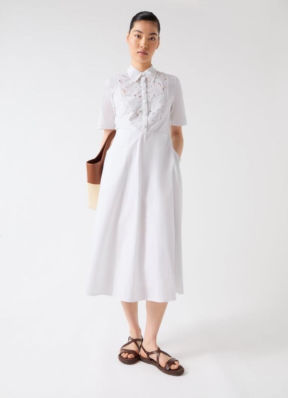This Grace dress is a timeless piece in white cotton poplin, featuring an embroidered lace bib panel, self-covered ball buttons, and pockets. It offers a sophisticated yet playful aura, ideal for smart casual events like garden parties or brunch outings. Pair it with strappy sandals or stylish ankle boots for a look that turns heads.