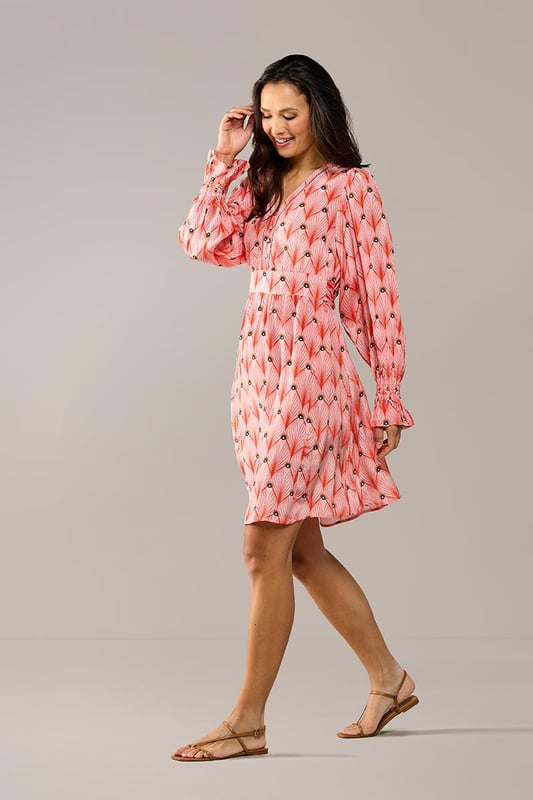 De elegante Belfast print tuniek uit de nieuwste collectie van Caroline Tensen is dé ideale keuze voor de zomer. De soepelvallende, wijde pasvorm en verfijnde details geven de tuniek zowel een chique als een ontspannen uitstraling. Het opvallende oranje pauwenprint op een zachte koraalroze achtergrond maakt deze lange tuniek bijzonder uniek. De lange mouwen hebben een subtiel ballon-effect en eindigen in speelse ruches bij de polsen, afgewerkt met elastiek voor extra comfort. De tuniek heeft een V-hals met een opengewerkte bies en een recht silhouet, waardoor hij heerlijk comfortabel zit. Dankzij het vaste strikceintuur kun je de taille mooi benadrukk