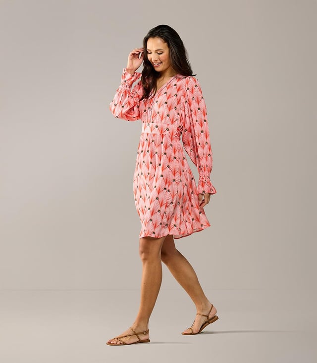 De elegante Belfast print tuniek uit de nieuwste collectie van Caroline Tensen is dé ideale keuze voor de zomer. De soepelvallende, wijde pasvorm en verfijnde details geven de tuniek zowel een chique als een ontspannen uitstraling. Het opvallende oranje pauwenprint op een zachte koraalroze achtergrond maakt deze lange tuniek bijzonder uniek. De lange mouwen hebben een subtiel ballon-effect en eindigen in speelse ruches bij de polsen, afgewerkt met elastiek voor extra comfort. De tuniek heeft een V-hals met een opengewerkte bies en een recht silhouet, waardoor hij heerlijk comfortabel zit. Dankzij het vaste strikceintuur kun je de taille mooi benadrukk