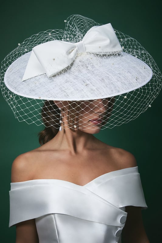 Striking wide brim fascinator with delicate pearl-adorned veil for a romantic finish
        Statement bow detail adds a contemporary touch to this classic bridal accessory
        Textured fabric base provides structure and sophistication
        Lightweight design ensures comfort throughout your special day
        Secures easily with discreet attachments for worry-free wear
    
    Make a captivating entrance on your wedding day with this exquisite Lisa Tan bridal fascinator from Coast. The wide brim silhouette creates a dramatic yet elegant profile, whilst the delicate veil adorned with subtle pearls adds a touch of romance and tradition. The statement bow detail introduces a modern twist to this timeless accessory, perfectly complementing an off-shoulder wedding gown as shown. This versatile piece works beautifully with both classic updos and flowing locks, allowing you to create your desired bridal aesthetic. Beyond the ceremony, this fascinator transitions seamlessly to the reception, maintaining its graceful presence throughout your celebrations. Pair with minimal jewellery to let this showstopping headpiece command attention, or match with pearl accessories for a coordinated bridal ensemble that reflects sophisticated British wedding style.
