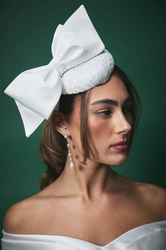 Elegant pillbox fascinator featuring a statement oversized bow detail
        Delicate lace detailing with subtle sequin embellishment for a touch of sparkle
        Secure headband design ensures comfortable wear throughout your special day
        Handcrafted from lightweight Abaca material for all-day comfort
        Versatile positioning allows for personalised styling to complement your bridal look
    
    Make a sophisticated statement on your wedding day with this exquisite Lisa Tan bridal fascinator. The classic pillbox silhouette is beautifully modernised with an eye-catching bow that adds dramatic flair without overwhelming your bridal ensemble. The intricate lace detailing creates a textural contrast against the smooth bow, offering a perfect balance of traditional craftsmanship and contemporary design. Style with an off-shoulder wedding gown as shown for a harmonious bridal aesthetic, or pair with a sleek updo to showcase dangling earrings for added glamour. This fascinator works beautifully with both classic and modern bridal styles, offering the perfect finishing touch whether you're walking down the aisle, posing for photographs, or celebrating at your reception. The secure fit ensures you can dance the night away with confidence.