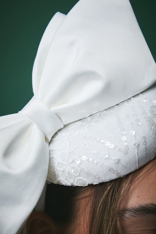 Elegant pillbox fascinator featuring a statement oversized bow detail
        Delicate lace detailing with subtle sequin embellishment for a touch of sparkle
        Secure headband design ensures comfortable wear throughout your special day
        Handcrafted from lightweight Abaca material for all-day comfort
        Versatile positioning allows for personalised styling to complement your bridal look
    
    Make a sophisticated statement on your wedding day with this exquisite Lisa Tan bridal fascinator. The classic pillbox silhouette is beautifully modernised with an eye-catching bow that adds dramatic flair without overwhelming your bridal ensemble. The intricate lace detailing creates a textural contrast against the smooth bow, offering a perfect balance of traditional craftsmanship and contemporary design. Style with an off-shoulder wedding gown as shown for a harmonious bridal aesthetic, or pair with a sleek updo to showcase dangling earrings for added glamour. This fascinator works beautifully with both classic and modern bridal styles, offering the perfect finishing touch whether you're walking down the aisle, posing for photographs, or celebrating at your reception. The secure fit ensures you can dance the night away with confidence.