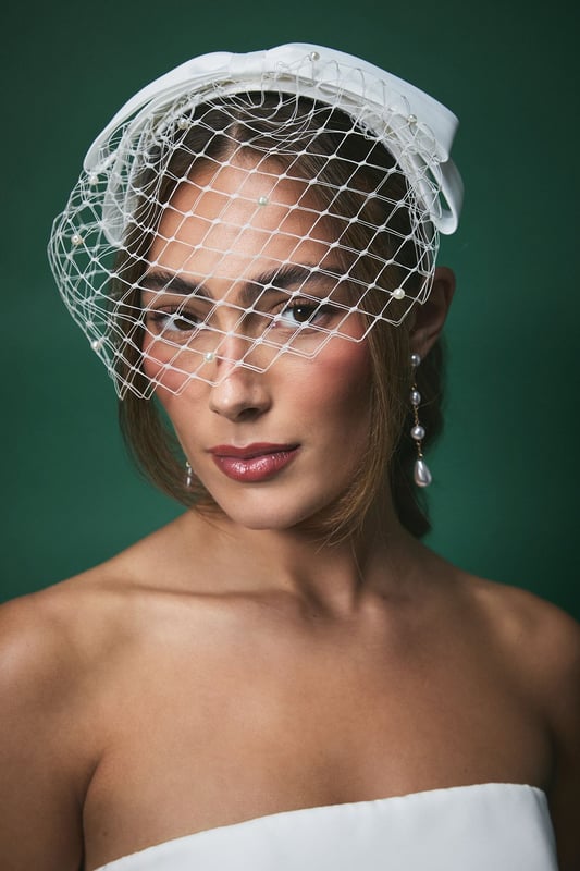 Elegant satin bow headband with delicate pearl-adorned veiling for a timeless bridal look
        Sophisticated birdcage veil design that frames the face beautifully
        Versatile one-size fit suitable for various hairstyles
        Intricate netting detail with subtle pearl embellishments throughout
        Structured bow accent that adds a touch of classic romance
    
    The Lisa Tan Bridal Bow Headband combines vintage charm with contemporary elegance, making it the perfect finishing touch for your wedding day ensemble. The delicate pearl-adorned veiling creates a soft, romantic frame for your face whilst the structured satin bow adds a touch of refined sophistication. This versatile headpiece complements both traditional and modern bridal styles, working beautifully with updos, loose waves, or sleek straight hair. Pair with simple drop earrings as shown for a coordinated look that won't overwhelm your bridal attire. The birdcage veil element offers a hint of mystery and allure, whilst still allowing your makeup to shine through. This headpiece strikes the perfect balance between statement and subtlety, ensuring you feel every bit the elegant bride on your special day.