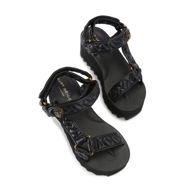 This Kurt Geiger Orion Pump sandal sits on a chunky black sole with exaggerated tread. The upper is in a black overstitch quilted leather alternative. The straps are fastened with velcro topped with an antiqued brass eagle head. Sole height: 7cm.