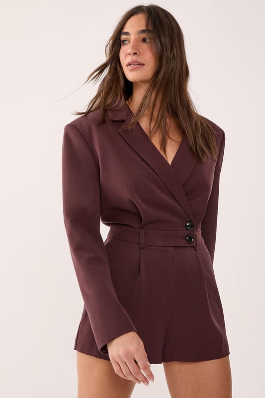 Tailored romper featuring a sophisticated wrap-style front with button detail closure
        Structured blazer-inspired lapel collar adds a smart, professional edge
        Long sleeves with subtle button cuffs for refined detailing
        Defined waistline creates a flattering silhouette
        Fully lined for comfort and smooth wear
    
    Make a statement in this striking tailored romper that seamlessly blends sophistication with contemporary style. The blazer-inspired design with its structured lapels and wrap-front detail creates a polished look that commands attention. Perfect for smart casual events where you want to showcase your fashion-forward sensibilities whilst maintaining an air of refinement. Style with knee-high boots for an evening out, or dress it down with chunky trainers for a modern daytime ensemble. For more formal occasions, pair with strappy heels and statement jewellery to enhance its elegant appeal. The versatile design transitions effortlessly from office to evening drinks, making it an essential addition to your wardrobe. This NastyGal piece offers the perfect balance of comfort and style for the fashion-conscious woman who isn't afraid to stand out.