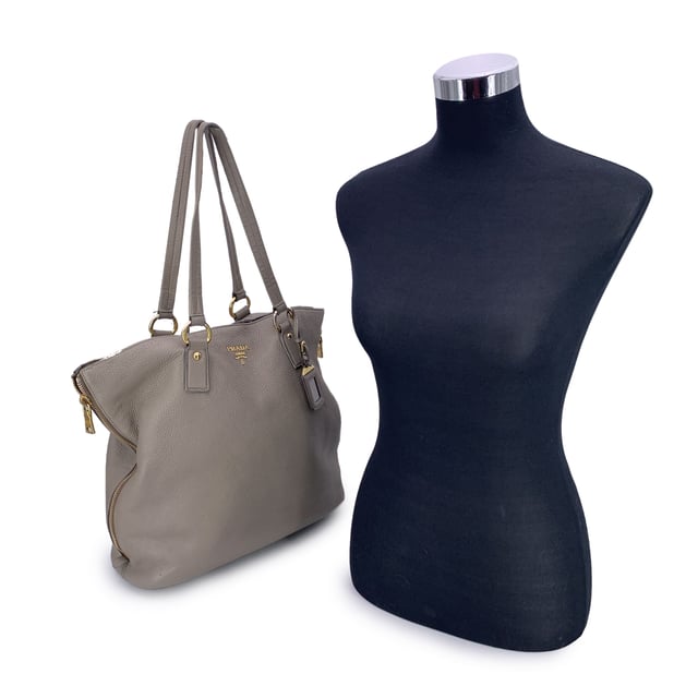 VINTAGE, RRP AS NEW
This beautiful item will come with a Certificate of ity provided by Entrupy. The certificate will be provided at no further cost  Elevate your everyday with this beautiful PRADA tote bag, crafted in luxurious grey taupe textured leather-a timeless neutral that pairs effortlessly with any look. Showcasing PRADA's signature blend of elegance and functionality, this tote features structured double top handles and refined silver-tone Prada lettering on the front, adding a touch of quiet sophistication.  The top opens with a button closure to reveal a spacious main compartment, fully lined in PRADA's iconic jacquard fabric. Inside, you'll find one zip pocket and one open pocket to keep your essentials neatly organized. A 'PRADA Milano' tag and a discreet numbered label tucked inside the interior pocket ensure ity.  Finished with four metal bottom feet for added protection and polish, this tote is as practical as it is chic-perfect for the modern woman who values understated luxury. Condition Rating: B - VERY GOOD CONDITIONCondition Details: Some wear of use on the upper border (please see photos). Some wear of use on the hardware. Please check the photos carefully and ask if you need to know more details or if you have any doubts.Measurements (cm):Depth: 15.2Height: 34.3Length: 36.8Other Details: Dustbag? NoKeys? NoBox? No Measurments:  width: 152 mm length: 368 mm height: 343 mm strap length: 0 mm handle drop: 0 mm