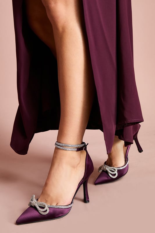 Elegant satin court shoes featuring a striking diamante bow detail at the toe
        Slender ankle strap with adjustable buckle fastening for a secure fit
        Pointed toe silhouette for a sophisticated, elongating effect
        High stiletto heel measuring between 7-9cm for a glamorous lift
        Versatile design perfect for formal occasions and evening events
    
    The Krissy Satin Diamante Bow Court Shoes manifest timeless elegance with a contemporary twist. The luxurious satin finish creates a refined canvas for the show-stopping diamante bow that adorns the pointed toe, catching the light with every step. Designed with a high stiletto heel that measures between 7-9cm, these shoes offer the perfect balance of height and wearability for special occasions. Style them with a tailored midi dress for weddings or pair with a sleek jumpsuit for race day sophistication. The delicate ankle strap not only provides additional support but also adds a feminine touch to the silhouette. These Coast heels are the ultimate finishing touch for any formal ensemble, whether you're attending a graduation ceremony, black-tie event, or celebrating a milestone occasion. Their refined aesthetic ensures they'll remain a treasured addition to your occasion wardrobe for years to come.